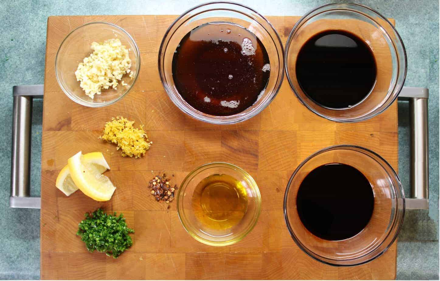 various small bowls filled with maple surop, oyster sauce, balsamic vinegar, garlic ,chilis and lemon zest on a wooden cutting board.