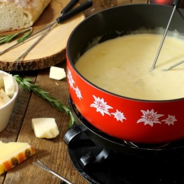 a red caquelon filled with real swiss cheese fondue on a wooden board surface surrounded by various ingredients