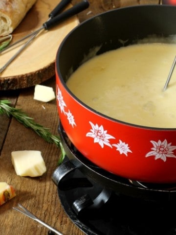 a red caquelon filled with real swiss cheese fondue on a wooden board surface surrounded by various ingredients