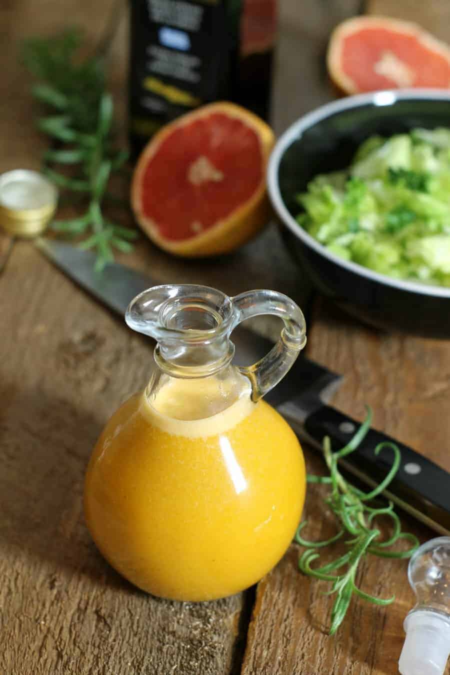 a glass bottle of grapefruit vinaigrette sitting on a wooden table top with a bowl of salad greens in the background.
