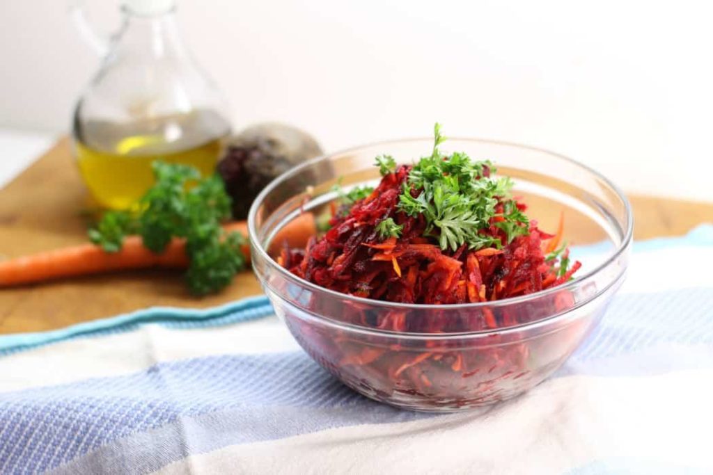 A glass bowl on a light blue dishcloth filled with a shredded beet salad with carrots and apples