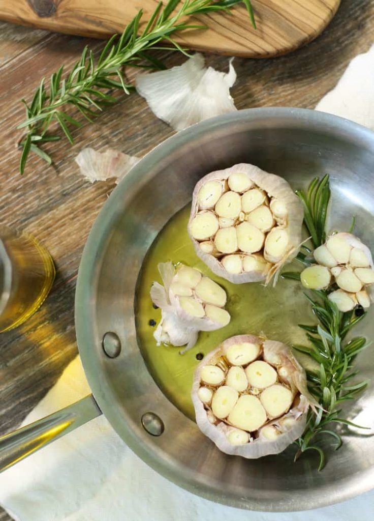 2 heads of garlic with the tops cut of and surrounded by herbs and oil, ready to be roasted in the oven