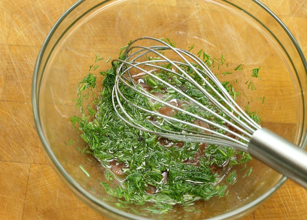 dill, vinegar, oil, and salt, being mixed in a glass bowl for the german cucumber salad dressing