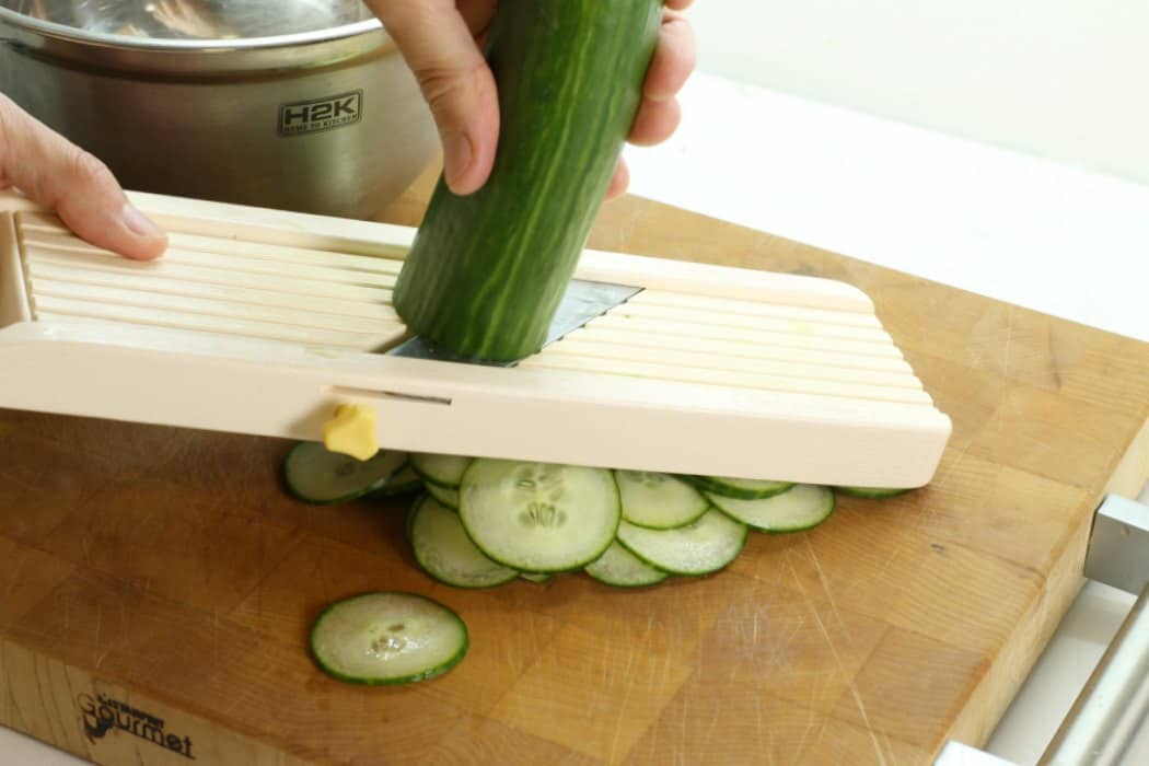 An english cucumber being sliced on a japanese mandolin slicer for a german cucumber salad.