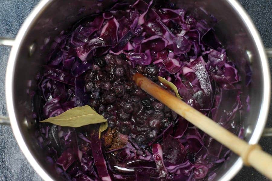 Chopped red cabbage cooking in a steel pot with blueberry jam and spices being added to the pot.