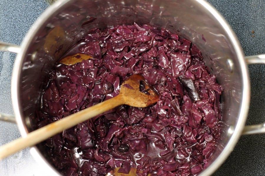 overhead shot of red cabbage braising in a steel pot with a wooden spoon in the pot about to stir it.