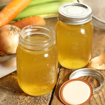 two mason jars on a wooden table filled with clear golden chicken stock. The jars are surrounded by various vegetables and a kitchen knife.