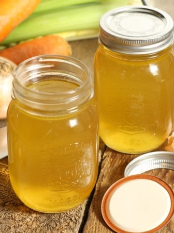 two mason jars on a wooden table filled with clear golden chicken stock. The jars are surrounded by various vegetables and a kitchen knife.