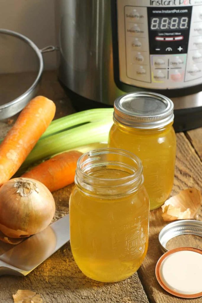 two mason jars filled with instant pot chicken stock sitting in front of an instant pot on a wooden tabletop.