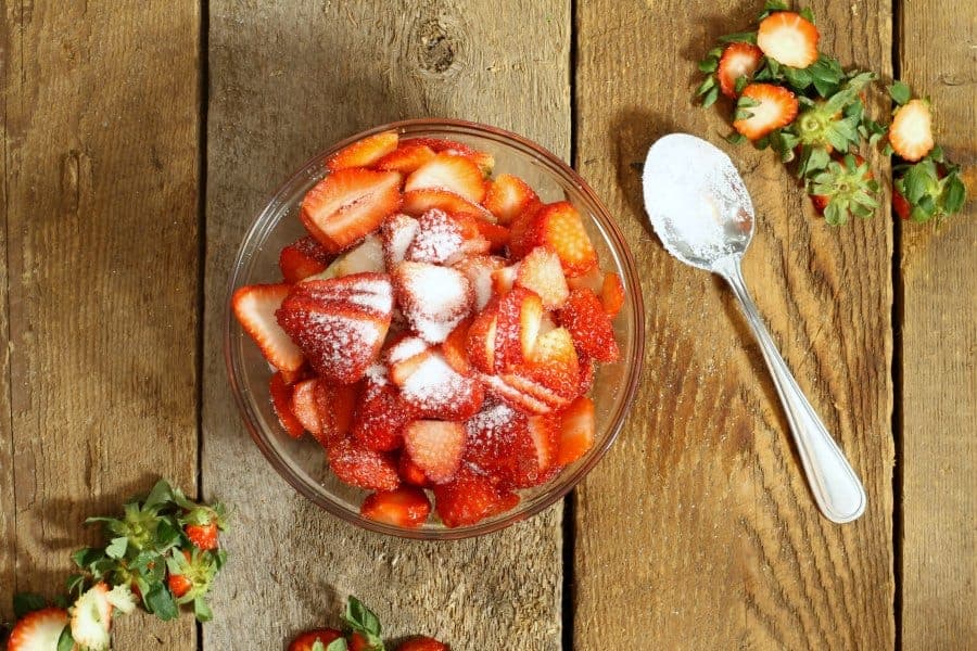A glass bowl sitting on a wooden table top, containing sliced strawberries sprinkled with white sugar. There is a spoon laying next to the bowl.