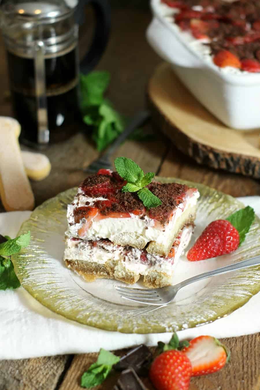 A piece of strawberry tiramisu sitton on a glass plate surrounded by a french press and strawberry and mint garnish