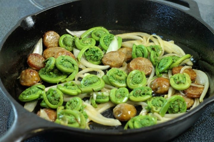 fiddleheads in a pan with italian sausages and onions in a cast iron pan