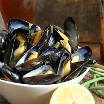 A bowl of beer steamed mussels with garlic scapes and lemon on a wooden table surrounded by various ingredients