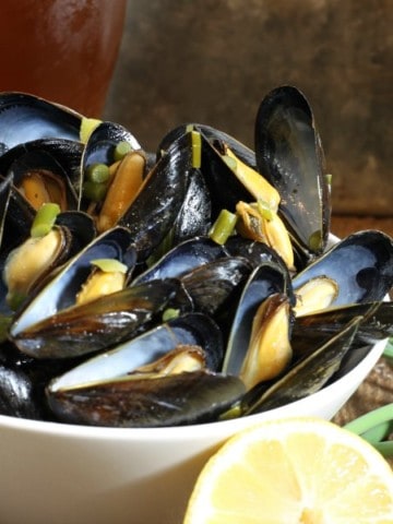 A bowl of beer steamed mussels with garlic scapes and lemon on a wooden table surrounded by various ingredients