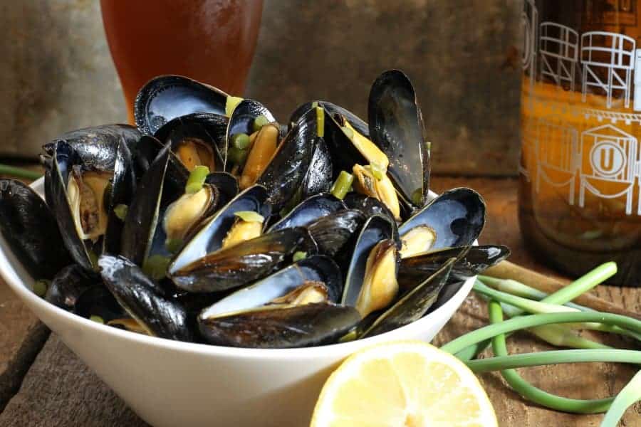 A bowl of beer steamed mussels with garlic scapes and lemon on a wooden table surrounded by various ingredients