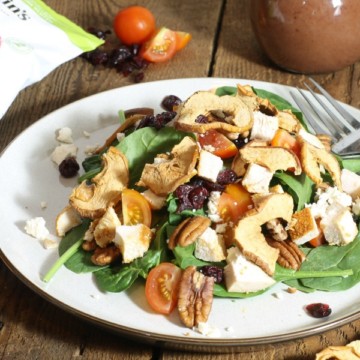 a chicken spinach salad garnished with crispy apple chips, feta, cherry tomatoes and pecans on a wooden table