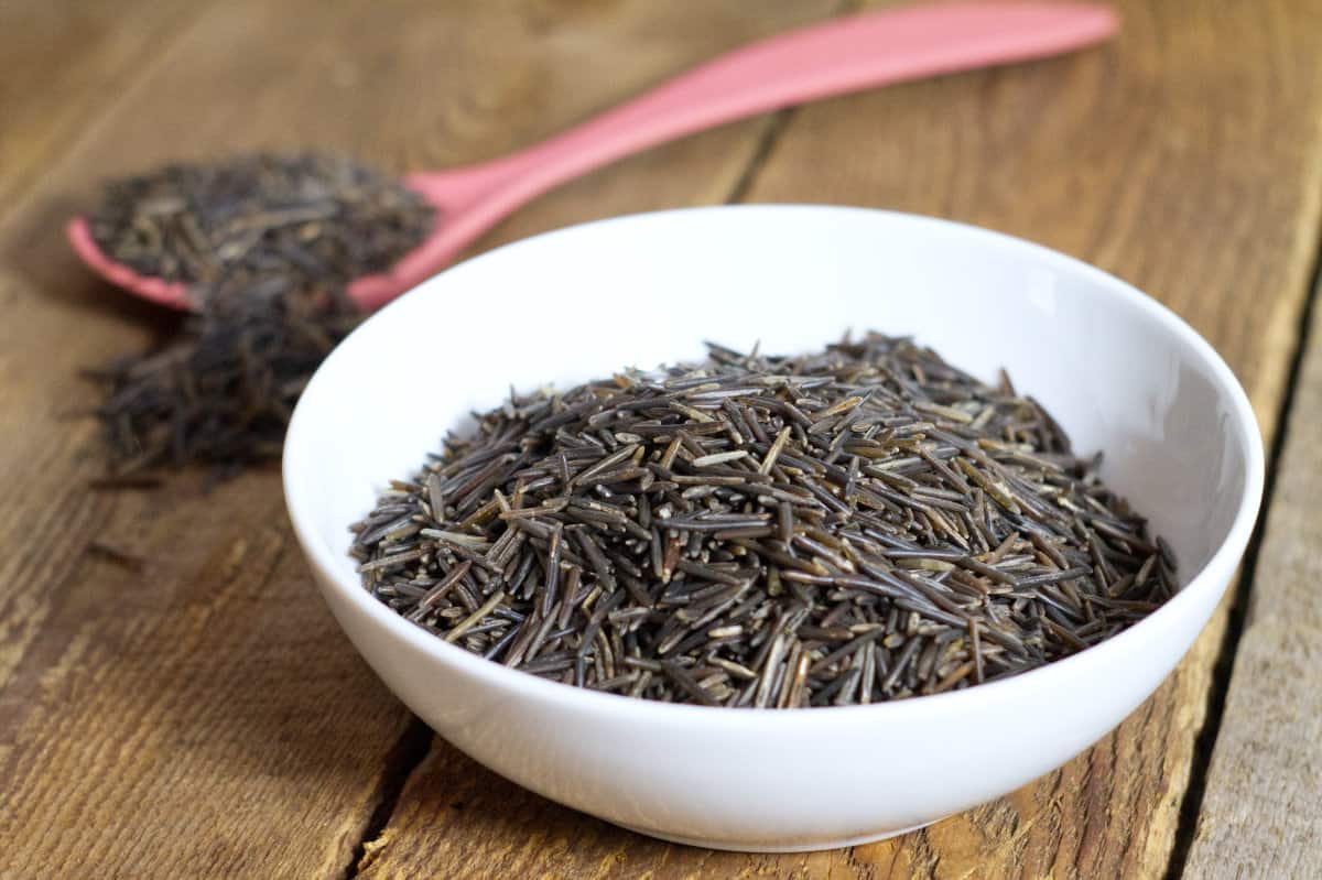 Raw Canadian wild rice in a white bowl, displayed on a rustic barn board table.