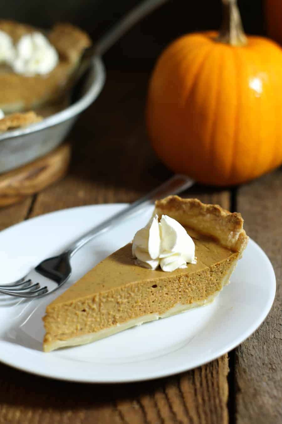 a single piece of homemade pumpkin pie on a white plate on a wooden table with a small pumpkin in the background