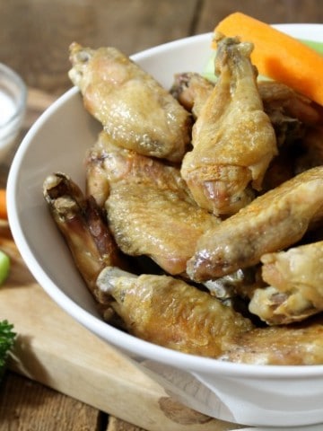 crispy baked chicken wings in a white bowl on a wooden tabletop with various garnishes.