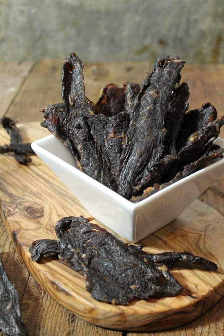 vertical image of oven dried beef jerky in a square white bowl on a wooden serving board