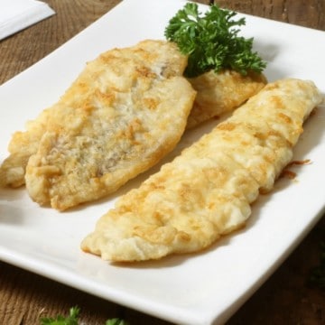 three pan fried pieces of haddock in a white plate with lemon wedges and parsley