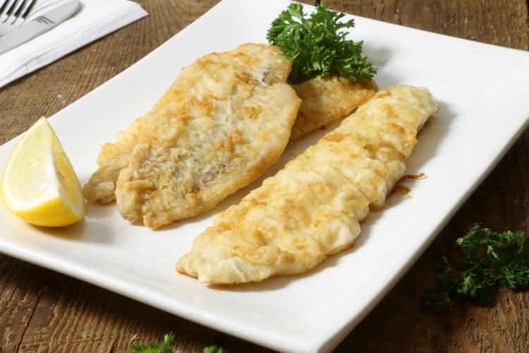 three pan fried pieces of haddock in a white plate with lemon wedges and parsley