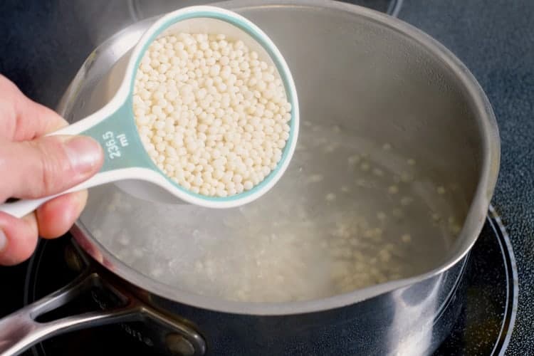 pearl couscous being added to a metal pot of boiling water.