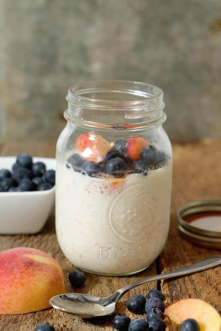 A glass mason jar on wooden barn boards filled with overnight oats with greek yogurt, blueberries and peaches