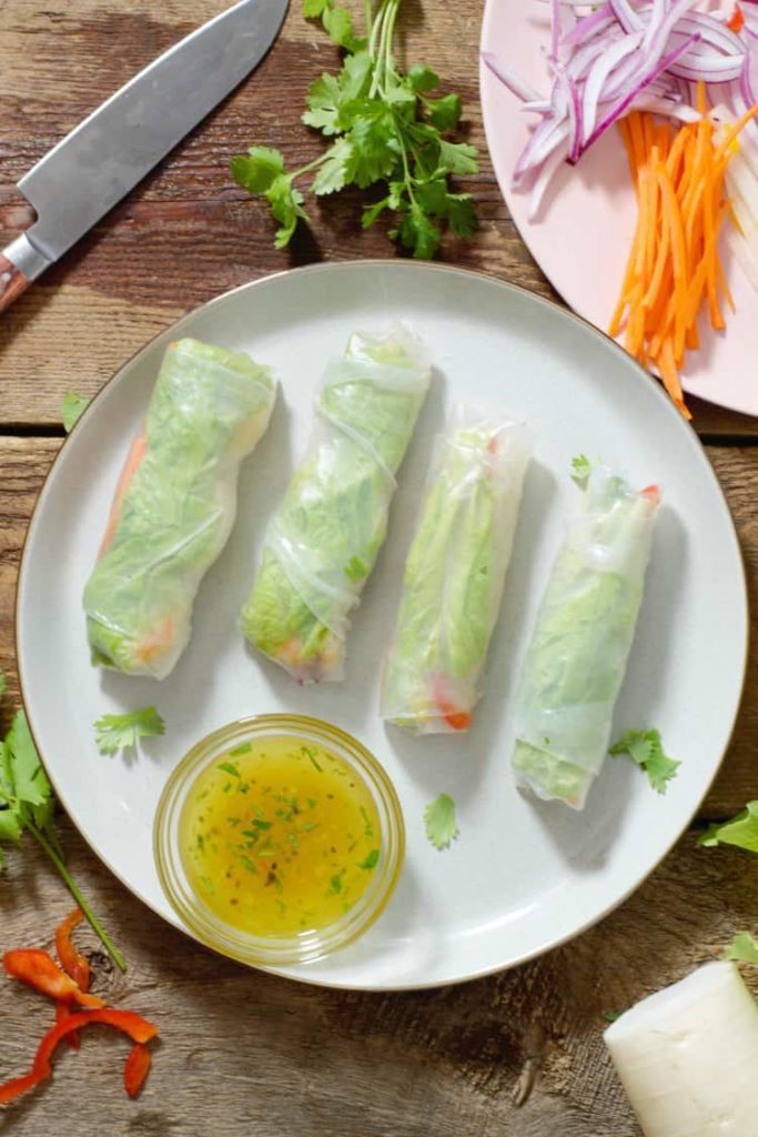 vertical image of rice paper rolls (vietnamese spring rolls) on a plate surrounded by dip and various ingredients