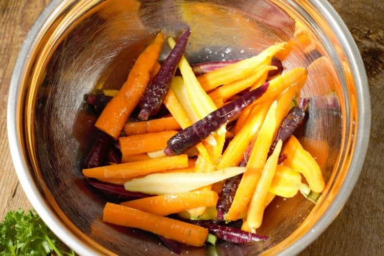 heirloom baby carrots being tossed in a metal bowl with oil and spices