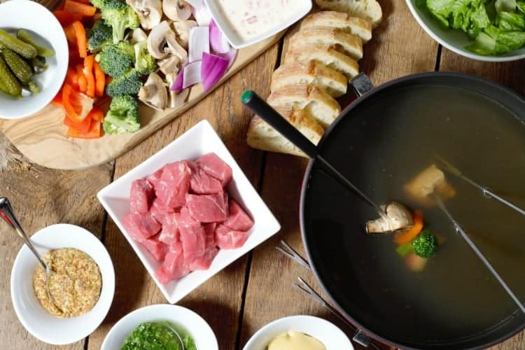 overhead shot of meat fondue being served on a wooden table fith raw veggies, dips, bread and more