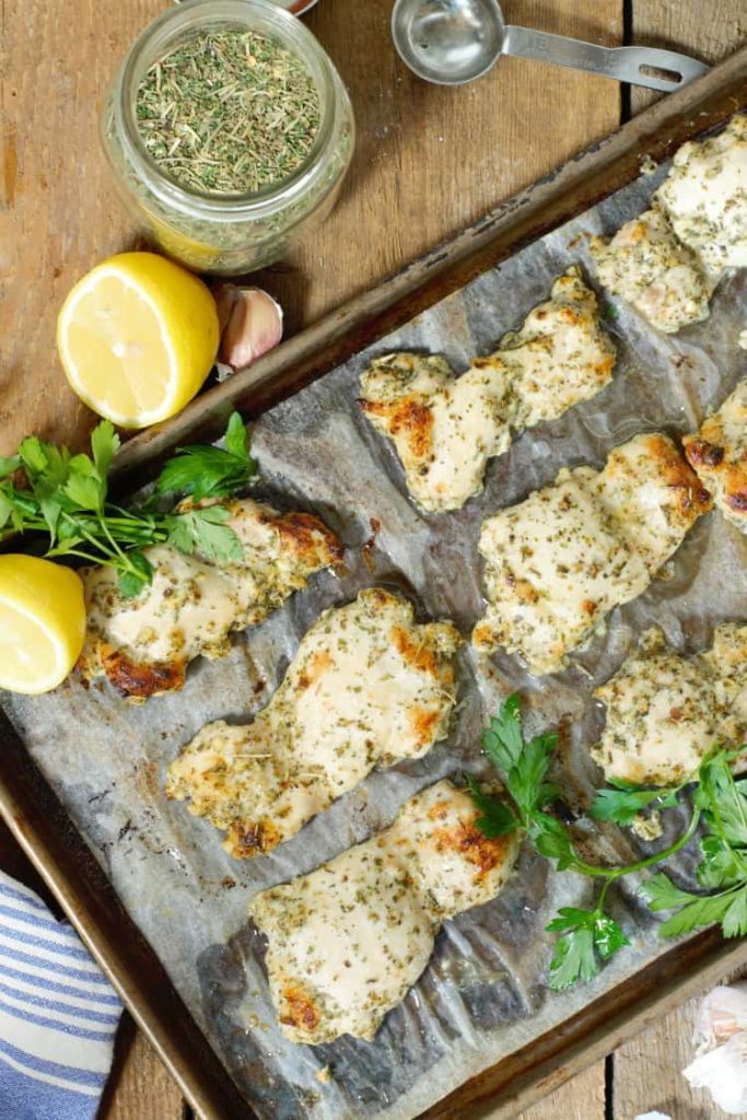 overhead shot of fully baked greek marinated chicken thighs on a grey sheet tray surrounded by various ingredients