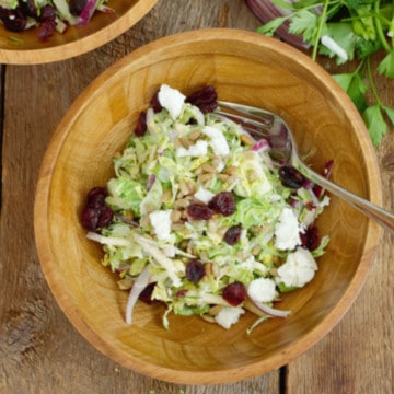 Overhead shot of shaved brussels sprout salad in a rustic wooden bowl, garnished with dried cranberries and goat cheese