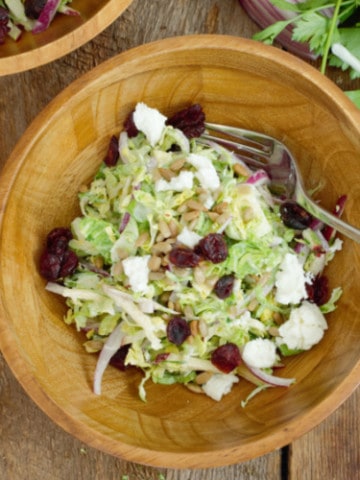 Overhead shot of shaved brussels sprout salad in a rustic wooden bowl, garnished with dried cranberries and goat cheese