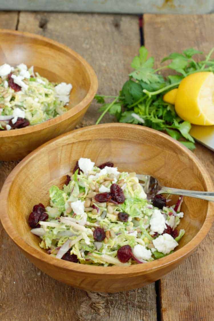 Vertical image of raw shaved brussels sprouts salad topped with goat cheese and dried cranberries served in a rustic wood bowl