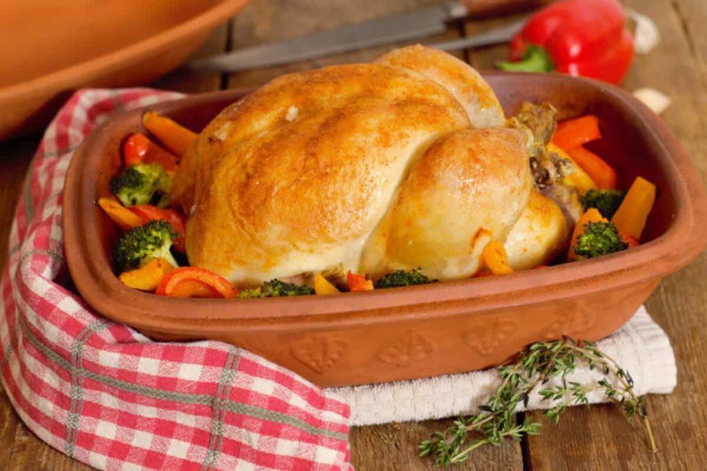 whole roasted chicken in a clay baker surrounded by bell peppers and broccoli