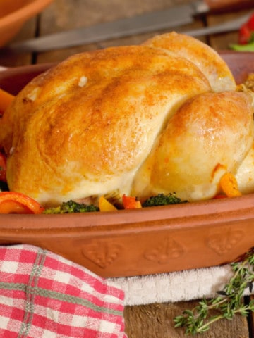 whole roasted chicken in a clay baker surrounded by bell peppers and broccoli