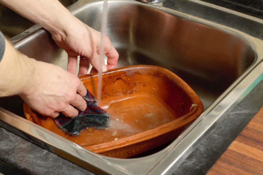 scrubbing a dirty clay baker with hot water to clean it