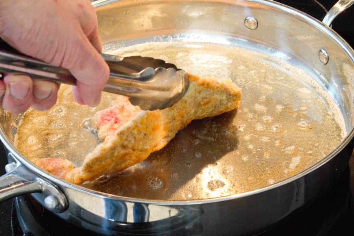 checking the underside of a partially cooked pork schnitzel with metal tongs to see if the pork schnitzel is ready to be flipped over