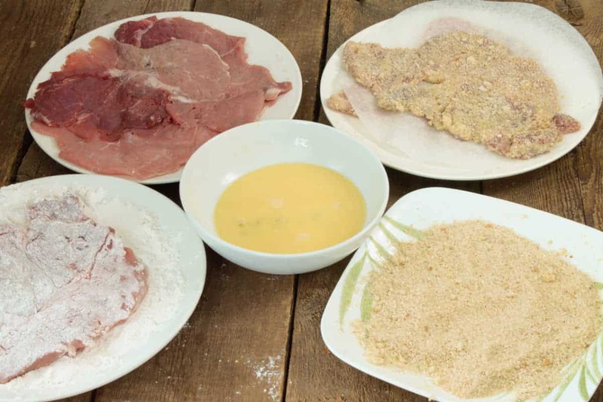 a breading station for making pork schnitzel consisting of four large plates. One with a flour mixture, one with a egg wash, one with a breading mixture and one with the raw pork cutlets.