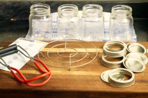 mason jars and lids, canning tongs, and other equipment displayed on a wooden cutting board