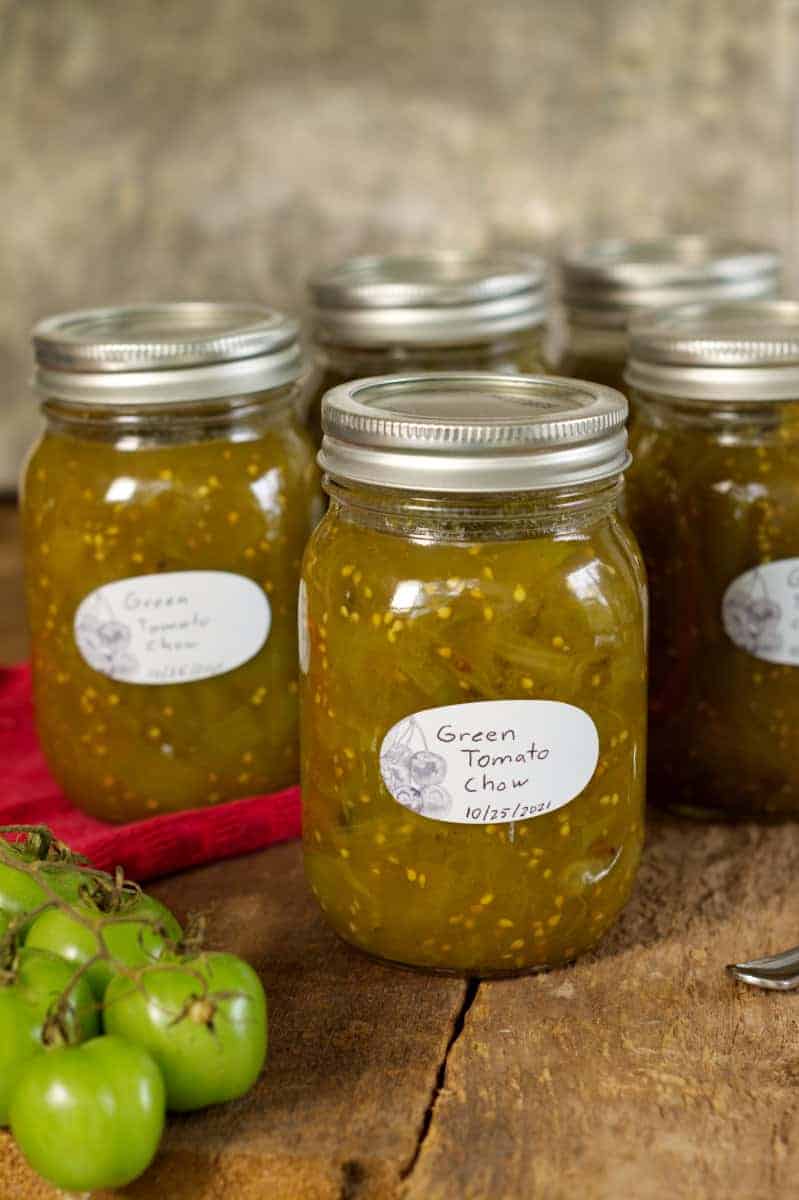vertical image of green tomato chow chow in labeled mason jars on a barn board surface.