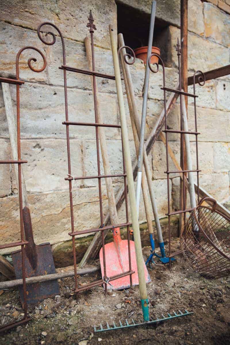 vertical image of various gardening tools leaning against an outside stone wall
