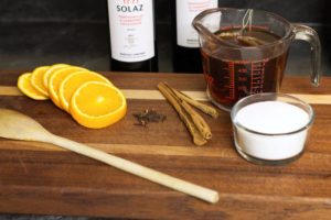 orange slices, a measuring cup of black tea, cinnamon sticks, and a dish of sugar displayed on a wooden cutting board