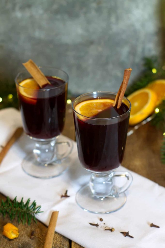 vertical image of two tall glass mugs filled with german gluhwein. the drinks are garnished with sticks of cinnamon and orange slices.
