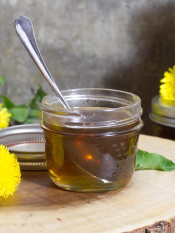 a small mason jar of dandelion syrup surrounded by freshly picked dandelion flowers
