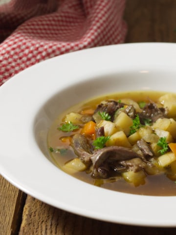 A close up of a white rimmed bowl filled with a broth based oxtail soup, displayed on a barn board background.