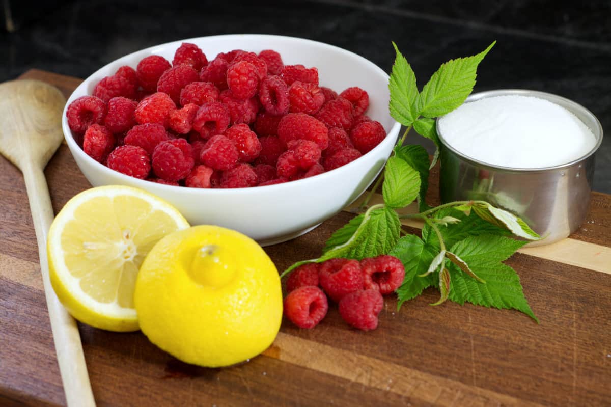 a white bowl of raspberries, a lemon cut in half, raspberry leaves, and a measuring cup of white sugar on a wooden cutting board.