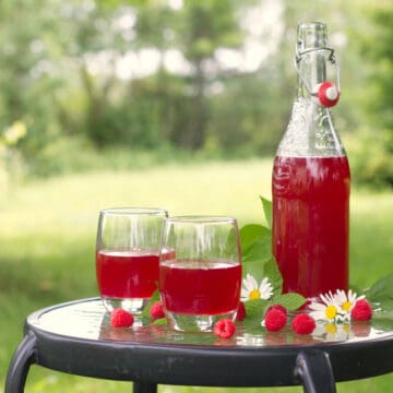 A glass bottle and two glasses of raspberry cordial served outside in a garden setting