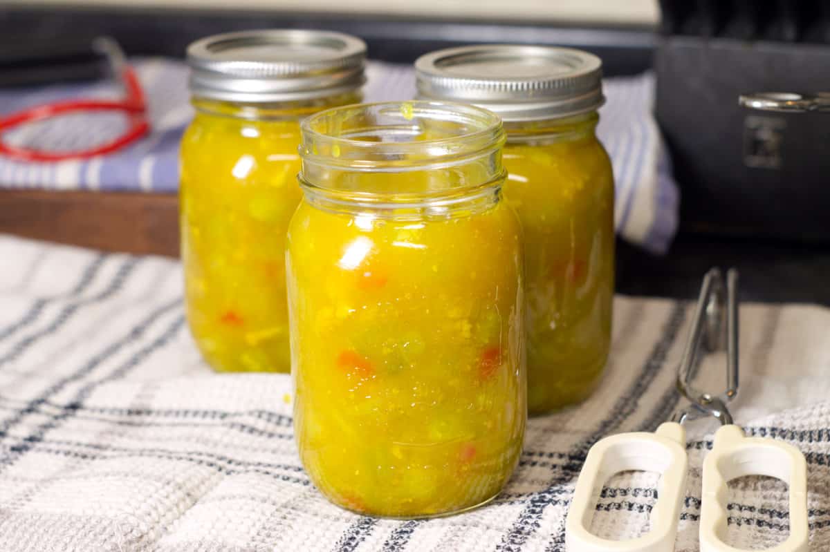 Three glass jars just filled with hot mustard pickles. the jars are displayed on a grey striped dish towel.